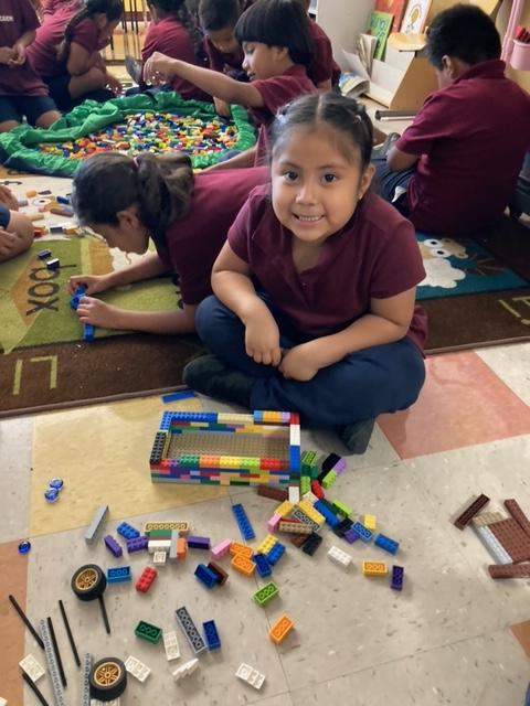 A student at Synergy Charter Academy smiles while sitting on the floor and building with LEGO bricks. She is wearing a maroon and blue school uniform. There are more students, also building with LEGO, behind her. 