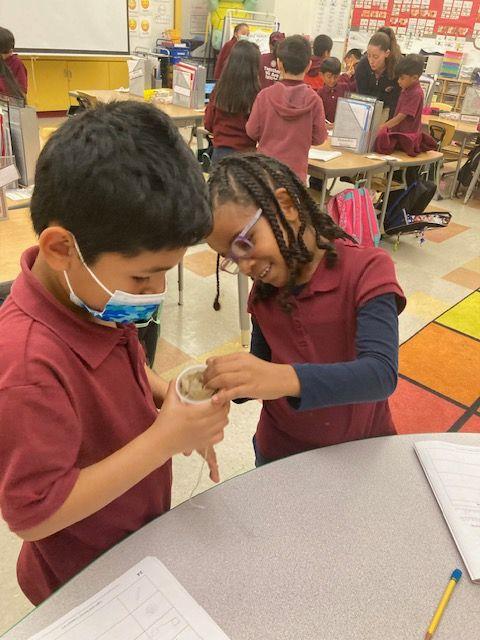  Two students at Synergy Charter Academy work together on a science experiment. They are standing near a table in a classroom and they are wearing maroon uniform shirts. There are teachers and students together in the background. 