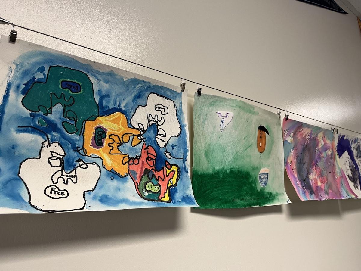 Student artwork hangs on a line in a classroom at Synergy Kinetic Academy. The brightly colored art is done with watercolors and black pen.
